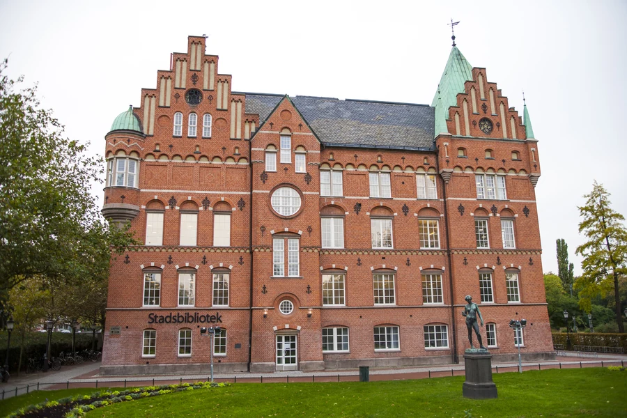 Fotografi av Malmö stadsbiblioteks äldre tegelbyggnad, med gröna koppartak och en grönskande park.